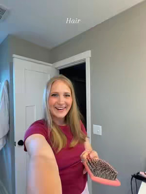 A selfie-style shot in a bathroom depicts a smiling woman who appears to be holding a hairbrush. The woman is blonde and dressed in a maroon t-shirt. The bathroom setting includes a white door, a mirror, and a light fixture. Above the woman's head is the word 'Hair'.