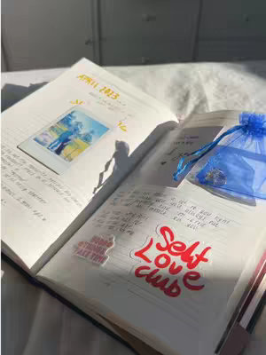 The image shows an open journal on a bed or similar soft surface, bathed in natural light. The left page has handwritten text and a photograph with an image of two people. The right side features more handwriting, a sticker reading 'Self Love Club' and a small blue bag with a charm.