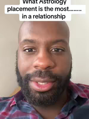 A close-up shot of a Black man with a shaved head and a full, dark beard. He is looking directly at the camera, and his lips are slightly parted as if speaking. He's wearing a plaid shirt, and the background is simple and out of focus.