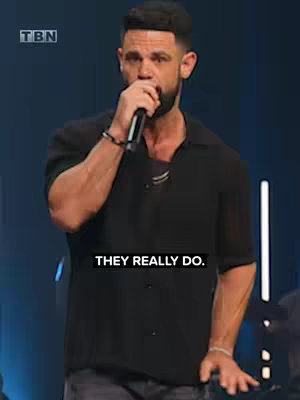 A man with short dark hair and a trimmed beard stands on a stage, holding a microphone. He wears a black button-down shirt and looks directly at the camera. The background is a dark blue with vertical, bright lights.