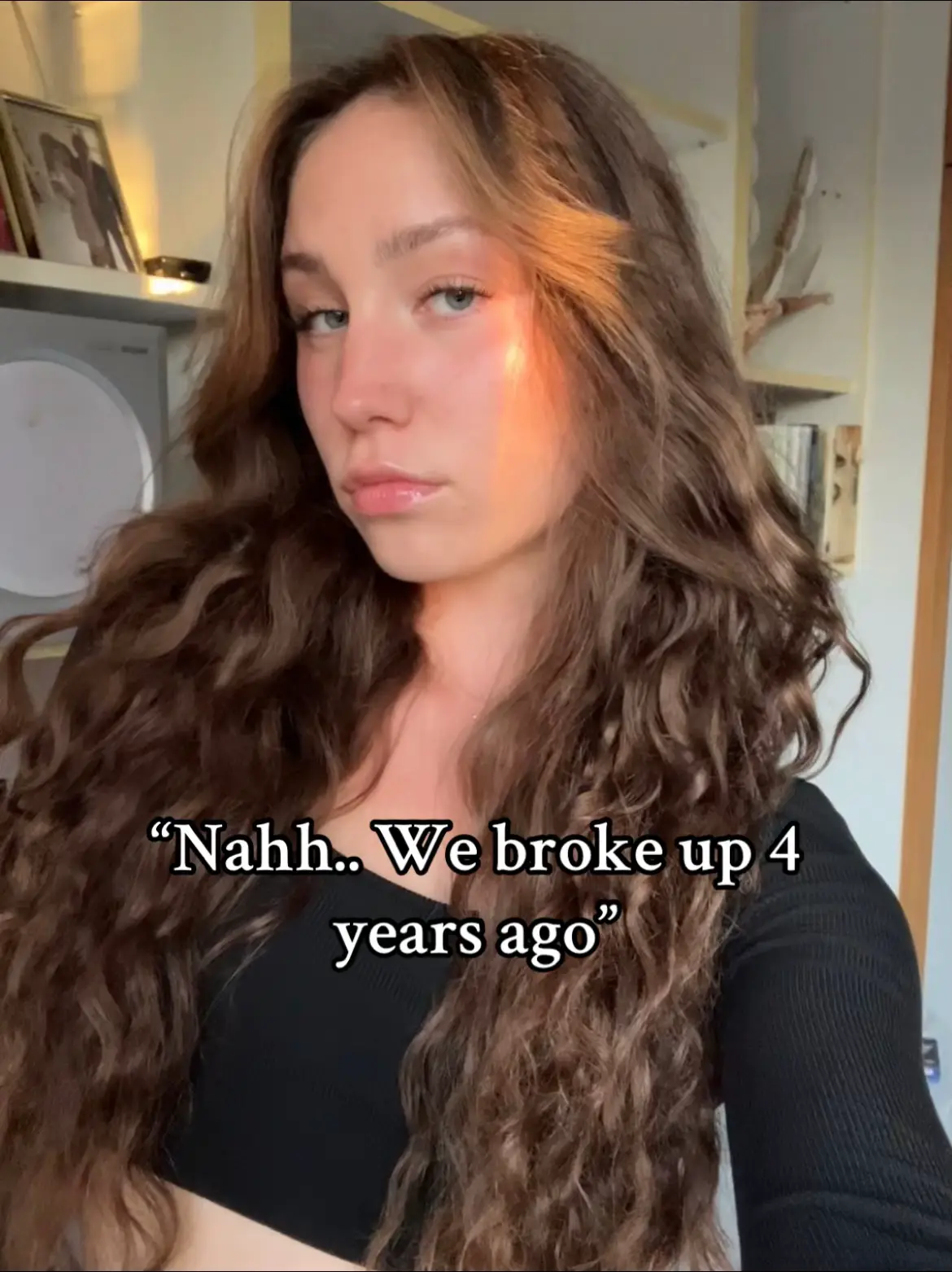 A woman with long, wavy brown hair is centered, gazing at the camera with a neutral expression. Text overlays are near the bottom, and there's a shallow depth of field suggesting a focus on the woman. Soft, warm lighting highlights the right side of her face.