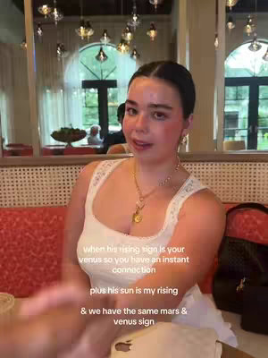 A woman with light skin and dark hair, wearing a white top, gold necklace, and earrings, sits in a restaurant with red seating and outdoor views. Text overlays are present on the video shot from the chest level.