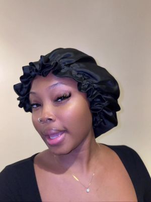 A young woman with dark skin is centered in the frame. She wears a black satin bonnet and a simple black top. Her expression is open, and she appears to be speaking. She has a nose ring and a delicate necklace. The background is a soft, neutral color.