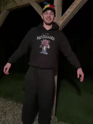 A man wearing a colorful hat and a black hoodie with an image on it stands with his arms outstretched in front of a barn structure at night. The scene is dimly lit.