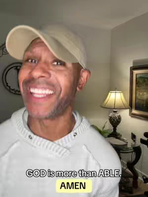 A man in a baseball cap and a white sweatshirt is smiling and looking at the camera. There's text on the image and an image of a framed picture and a lamp are visible in the background.