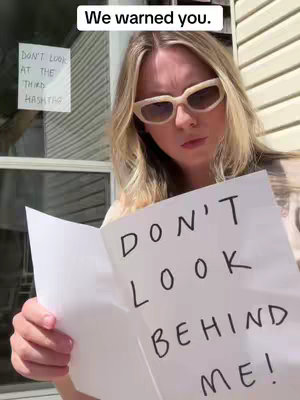 A woman wearing large cream-colored sunglasses holds up a white card where she writes 'Don't look behind me'. There is a sign on the glass behind her that says, 'Don't Look At The Third Hashtag'. In the top left corner, there is text that says 'We warned you.' This image is taken outdoors with a view of a paneled wall behind the woman.