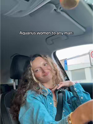 A selfie-style shot taken from inside a car, featuring a woman with highlighted brunette hair. She's wearing a denim jacket and gold accessories, including a watch and bracelet. The image is well-lit, and the woman has a slightly amused expression while looking straight at the camera, as if driving.