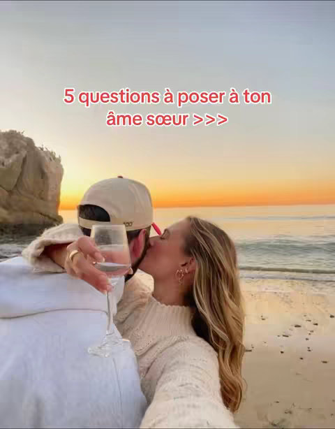 A couple embraces on a beach at sunset. The woman, facing the camera, kisses a man who is turned away, holding a glass of wine. Text overlayed reads,  "5 questions à poser à ton âme sœur >>>," in a red font.