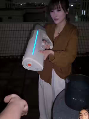 A woman is standing on a rooftop, holding a white device with a blue light; she appears to be demonstrating it to someone off-camera. The background is dark, and there are other rooftop objects, like a dark container, visible.