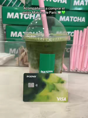 The image shows a close-up of a matcha drink in a clear plastic cup with a pink straw. In front of the drink is a Pixpay card and a green container that says “Matcha”. In the background are more green boxes and pink straws.