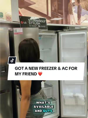 A person, viewed from the back, is looking at home appliances inside a store. The person is wearing a black top and jeans. In the view, there is a silver refrigerator and the open door of a refrigerator. The background shows a retail store with more appliances and signage.