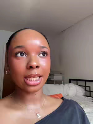 A close-up shot of a woman looking up and to the right, her face illuminated with bright light that gives her skin a reddish hue. Her lips are slightly parted, and she appears to be speaking. Her hair is pulled back. She wears silver hoop earrings and a silver necklace. The background is a blurry bedroom.