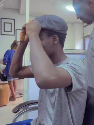 A person is adjusting a hat indoors, with another person partially visible in the background and a framed picture on the wall. The lighting is bright, and the scene is candid.