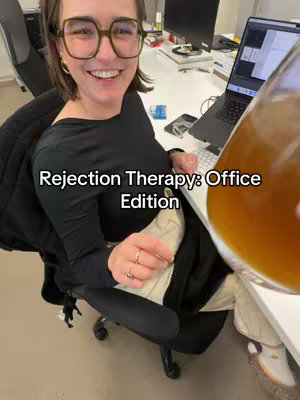 A woman in glasses is seated at a desk in an office setting. She is smiling and holding a clear glass filled with a light-brown liquid. Text overlay reads, 'Rejection Therapy: Office Edition.'