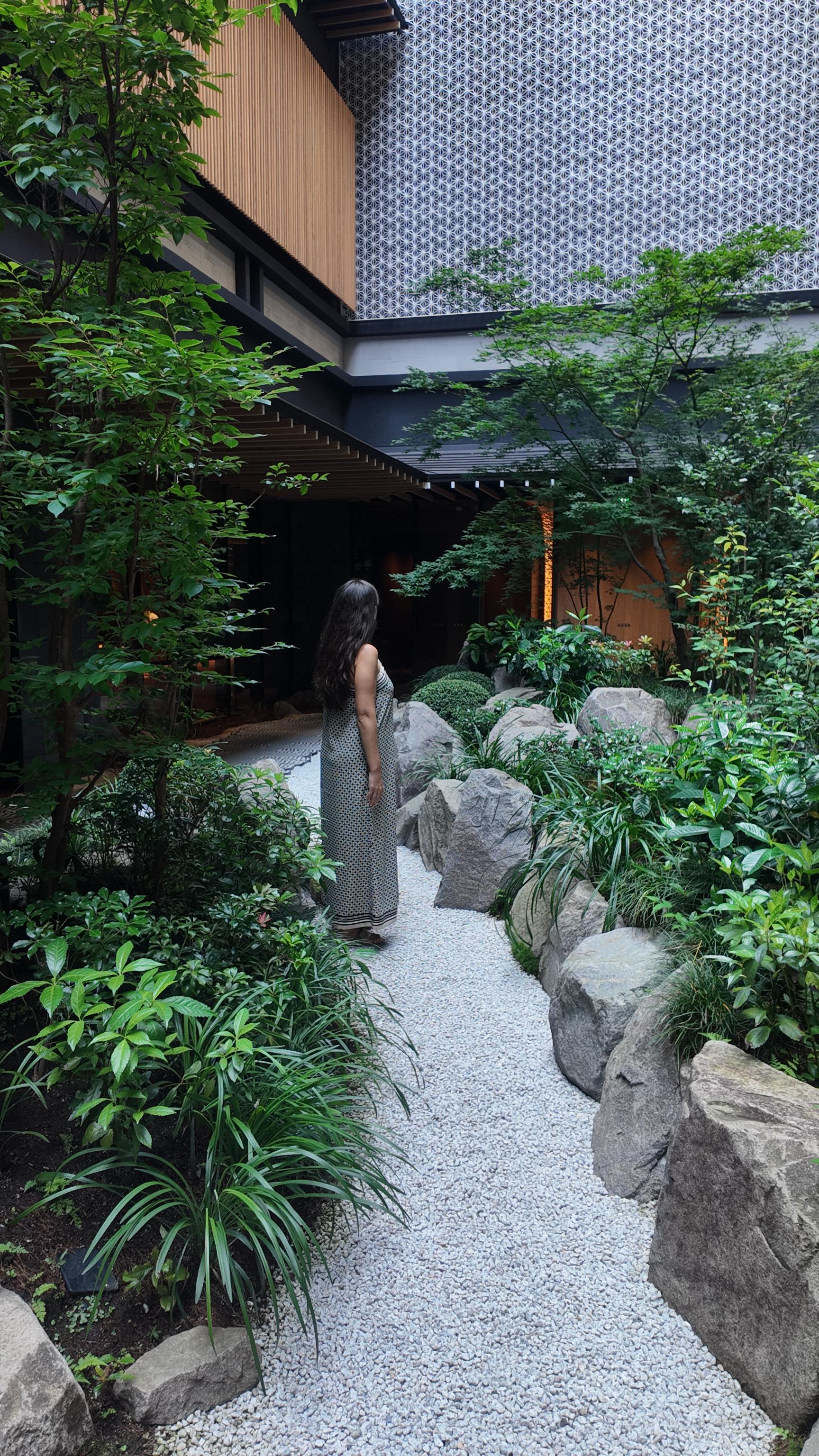 The image captures a serene outdoor scene within a Kyoto hotel. A person with long dark hair, seen from the back, stands on a white gravel path, looking towards a shaded area. Lush greenery surrounds the path, featuring various plants and large stones that create a natural, aesthetically pleasing environment. Architecture frames the scene, with wooden and patterned walls adding to the Japanese aesthetic.