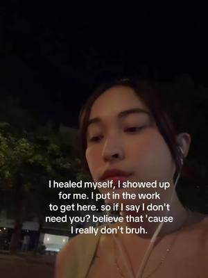 A medium shot of a young woman in a dark outdoor setting at night. The woman, with dark hair pulled back, is looking down and slightly to her left, with a serious expression. Dressed in a light-colored top, she has earbuds in. A series of text overlays are positioned below her face. The background is dark; trees are visible with blurry lights indicating a street.