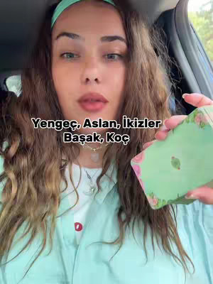A woman in a light blue button-up shirt and a green headband sits in a car, holding a deck of cards. Overlayed text lists zodiac signs. The woman has long brown wavy hair, and is looking towards the camera.