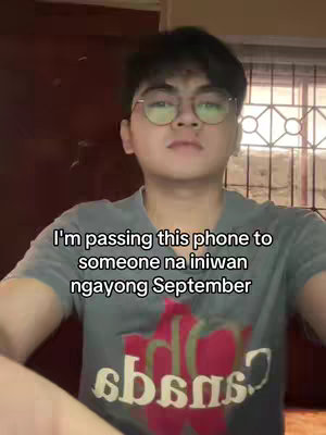 A young man with short black hair and glasses is centered in the frame. The background is a plain wall and a window with light coming through. He is wearing a gray t-shirt with a graphic on it. Text is overlaid on the image.