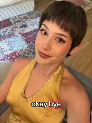 A selfie-style shot featuring a woman with short, dark hair and dark lipstick. She wears a yellow halter top with gold detailing and smiles slightly at the camera. The text "okay bye" is overlaid on the lower part of the image. The background is slightly blurred, suggesting a casual indoor setting.