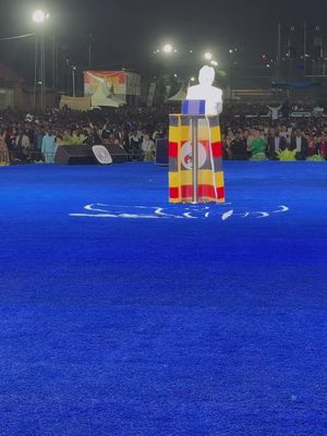 The image shows an outdoor scene at night with a large crowd gathered. In the foreground, a raised platform draped with the Ugandan flag features a translucent figure. The background is filled with blurred figures illuminated by lights, creating a sense of a large event or gathering.