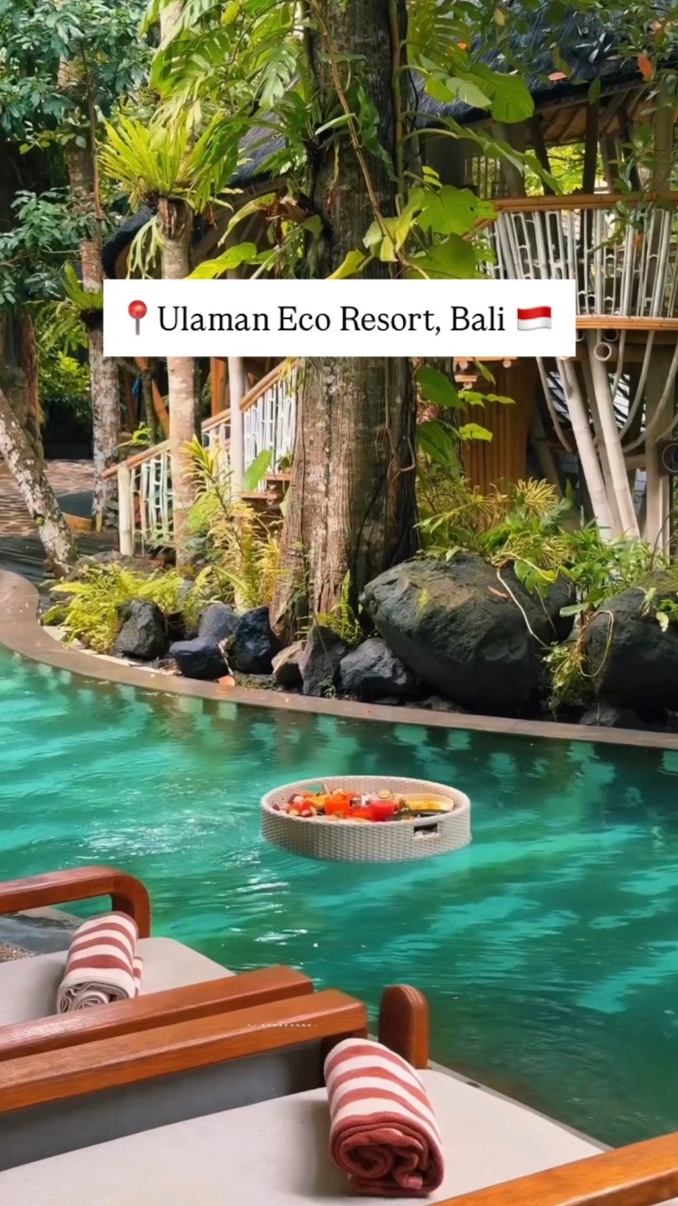 A serene scene featuring a pool of clear, turquoise water surrounded by lush green foliage, including trees and plants. A semi-submerged tree house and white railings with stairs along the pool add architectural elements. A floating tray laden with food is positioned in the water. The foreground shows two lounge chairs with rolled up towels, against a partly visible wooden deck. Text overlay appears in a white boxed, indicating the location pin. The overall composition is vibrant and evokes a sense of luxury and tranquility.