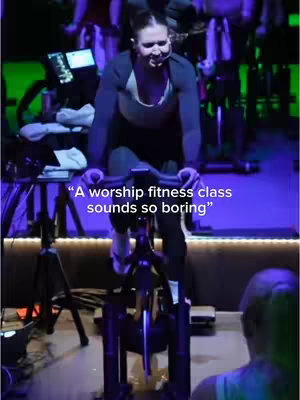 The cover image shows a woman on a stationary exercise bike inside a fitness studio. She is riding the bike, wearing a headset mic, and smiling. The background shows additional exercise bikes and people out of focus. Text on the image overlaid says "A worship fitness class sounds so boring".