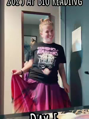 A woman stands in a room, smiling and holding up the edges of a flared pink and purple skirt. She wears a black t-shirt with a graphic and her hair is pulled up. The image includes a mirror and a partial door.