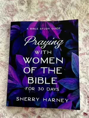 The image features a book cover with a floral background in shades of purple and dark blue. The book's title, "Praying with Women of the Bible for 30 Days," is prominently displayed in white and silver lettering, centered on the cover, with the author, Sherry Harney, listed at the bottom. The layout is vertical, and the overall composition suggests a focus on religious study and reflection.