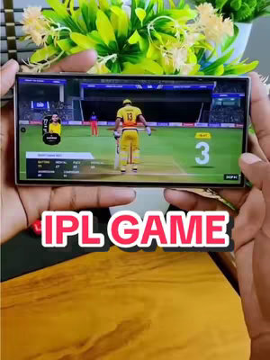 The cover image shows a person holding a smartphone with a cricket game displayed on the screen. The words "IPL GAME" are overlayed in red and white text. The background of the shot includes some green and yellow decorative plants.