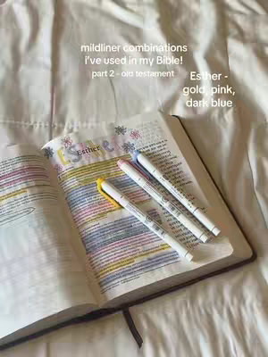 The image shows an open Bible on a bed and several highlighters. The Bible is open to the book of Esther, with the text highlighted in yellow, pink, and blue. Three highlighters are laid across the open book. The bedding is white. The layout is fairly simple, providing a focused view of the Bible, highlighters, and their respective markings.