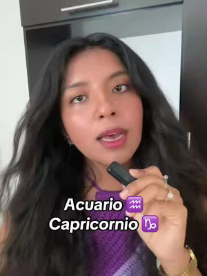 A woman with long dark hair faces the camera and speaks while holding a microphone. Text overlays include "Acuario Capricornio" and zodiac symbols. Behind her is a neutral background.