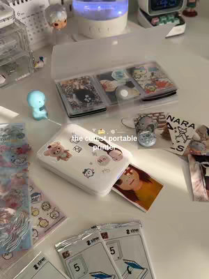 A well-lit photo presents a desk setup featuring a white portable photo printer with stickers, a small, clear case holding photos, and various desk accessories. The scene is styled with a kawaii aesthetic, with items neatly arranged on a white surface.