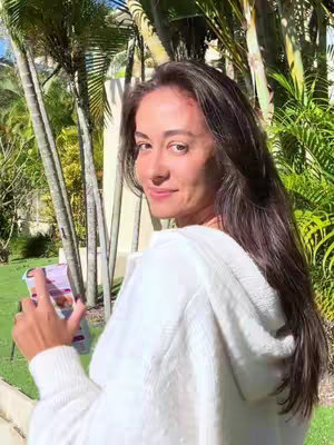 A woman with long, dark hair turns her head towards the camera, smiling. She's wearing a white hoodie and holding a purple product, set against a backdrop of green foliage and palm trees.