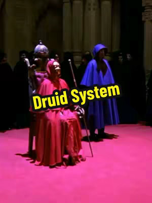 The image shows a scene with several figures in what appears to be a ceremonial or ritualistic setting. The foreground features two main figures: one is seated, wearing a red robe with a hood that obscures their face, and holding a staff. The staff looks like a magic wand. Beside this is a man in a blue robe standing. Behind them is an environment in a darkened hall or chamber, with other figures in the background, all in dark clothing, suggesting a gathering or ritual. The flooring is bright pink, and the overall lighting is dim. Overlaid on this scene is text in a yellow, outlined font that reads "Druid System."