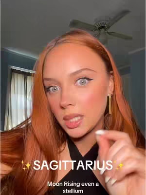 A selfie-style video presents a young woman with long auburn hair, fair skin, and striking blue-grey eyes. She is wearing a black top, gold earrings, and has a touch of blush and lip gloss. The background is slightly blurred, showing a room with a ceiling fan and window curtains.