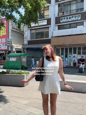 A woman stands in front of a building, wearing a white dress and engaging an open arms pose. The background includes commercial buildings, a green tent-like structure, and various signs, creating an urban environment.