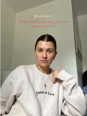A medium shot features a woman in a cream-colored sweatshirt with the words 'Jesus is Lord' visible on the chest. Light from an unseen source illuminates her face and upper body. She gazes directly at the camera with a calm expression, one hand near her face. Behind her, the wall is plain and off-white. A text overlay in pink, centered at the top, reads 'Jesus says: If the world hates you, just know it hated me first'.