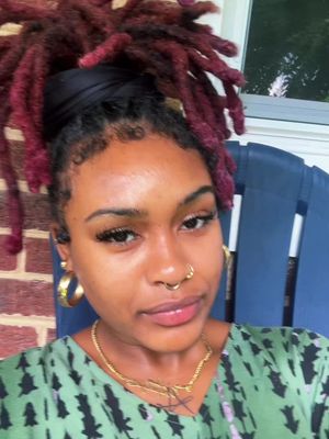 A close-up shot of a young woman, framed from the chest up. She has dark skin, large gold hoop earrings, a nose ring, and a gold necklace. Her hair is styled into red dreadlocks that are pulled up with a black head wrap. She is looking directly at the camera with a slight smile. She wears a green patterned top, and the background is a blurred outdoor setting, possibly a porch with brick and a blue chair. The lighting is natural and appears soft.