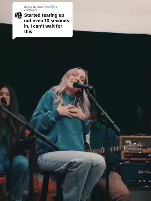 A woman with long blonde hair, wearing a teal hoodie and jeans, sits on a stool and sings into a microphone. A commenter's text overlay is in the top-left corner. Behind her are other band members, a microphone, speakers, and other equipment, on a dark background.