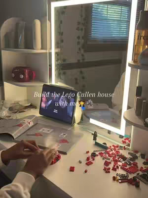 A close-up shot of a person's hands working on a red Lego build, with various Lego pieces scattered on a white surface. A tablet showing a video of a movie scene is set up at the rear of the workspace, next to a lit vanity mirror and other items. The overall composition is tidy and well-lit, with a focus on the Lego building process.