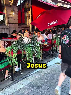 An outdoor scene at a restaurant with several people seated at tables under red umbrellas. Lush green plants and decorations line the sides of the seating area. A cobblestone walkway borders the scene, and a person in a black shirt is seen walking past the restaurant.