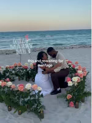 A man kneels, proposing to a woman on a beach. The woman is in a white dress. They are framed by a ring of flowers with candles, with the words 'Will you marry me?' shining in neon in the background.