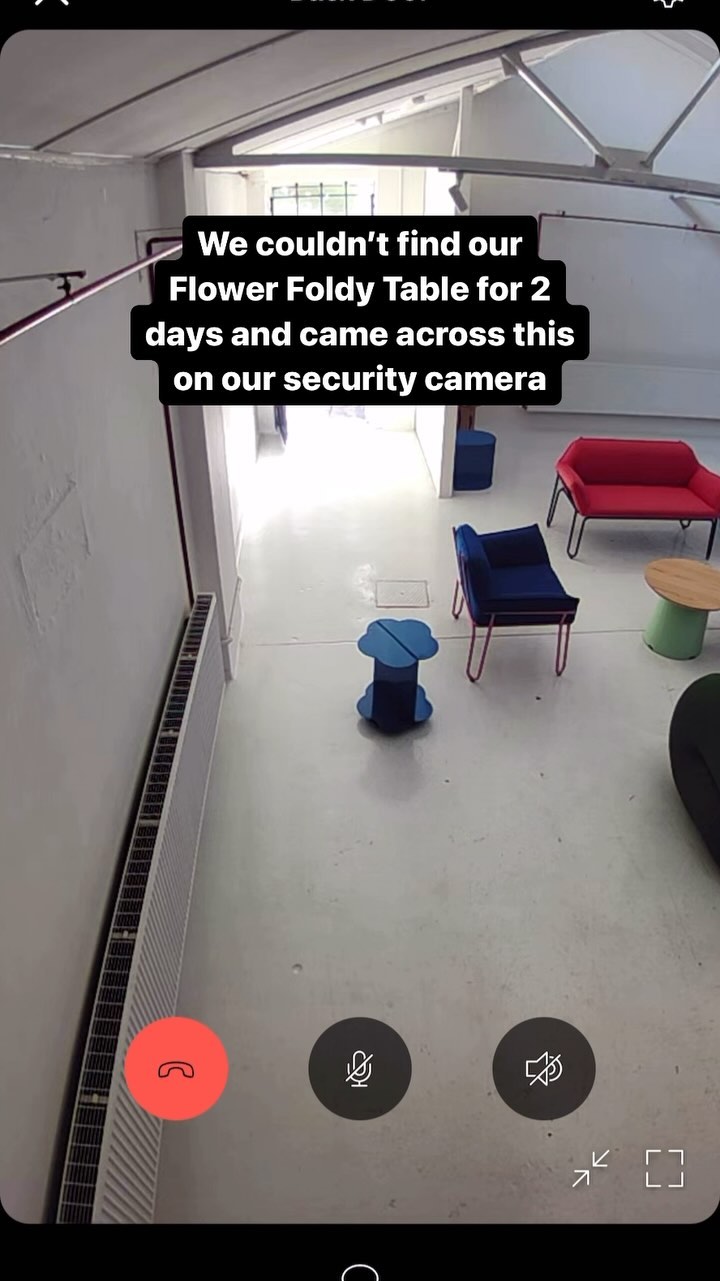 A still from a security camera, showing an empty room with modern furniture. The room is brightly lit with white walls and a shiny, reflective floor. A long, white radiator runs along one wall and the room has a glass door at the end leading to a hallway. The focus is on a blue flower-shaped table in the center of the room. The video call overlays display buttons to end or mute the call.