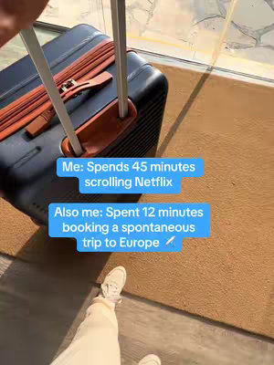 An overhead shot featuring a partially visible, dark blue travel suitcase with brown leather accents. The side of the suitcase is facing the camera, while the handle is extended upwards. Below the suitcase, a pair of light-colored legs and feet are visible, wearing white sneakers. The suitcase sits on a brown carpeted surface near a door or window. Two blue rectangular text boxes are overlaid on the image, containing text related to Netflix and Europe trip planning.