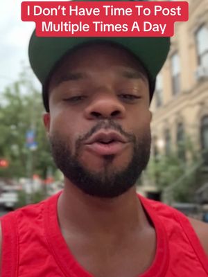A selfie-style shot with a man against a blurry outdoor background. The man, who appears to be the primary subject, is in the foreground, framed from the chest up. He's wearing a red tank top and a dark green baseball cap. Overlayed at the top of the image is red rectangular banner with white text that reads, "I Don't Have Time To Post Multiple Times A Day". The background seems to be a street scene with buildings and possibly some greenery, however, the focus is clearly on the person and this overlaid text. The man's expression is neutral, with his lips slightly parted. The lighting appears to be natural, and the overall composition is straightforward and casual.