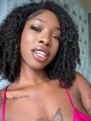 A close-up shot of a Black woman with curly hair looking slightly upwards and to the side with a neutral expression. She is wearing a pink top that reveals a tattoo on her chest and another located on her upper chest. Behind her is a blurred, light-colored headboard.