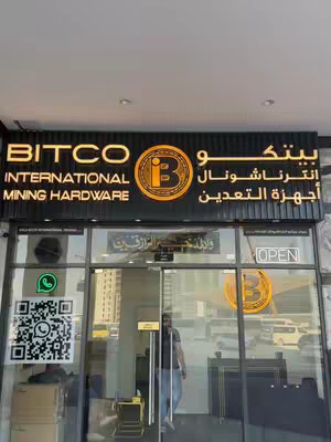 The image displays the exterior of a store. The store sign, prominently displayed above the entrance, features the name "BITCO INTERNATIONAL MINING HARDWARE" in bold, illuminated letters. The store's name is also written in Arabic script. The entrance is a glass door with a person partially visible inside. The windows reflect the street scene, adding depth to the image.