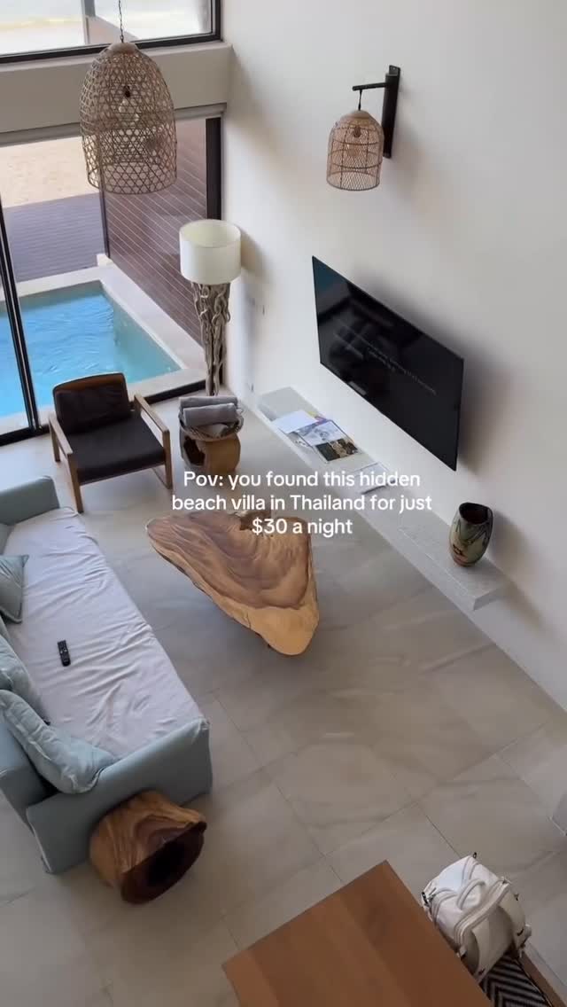 An overhead shot captures the interior of a modern beachfront villa. The scene showcases a light-blue couch in the foreground. A tree-trunk-shaped table sits prominently in the room’s center. The backdrop reveals a pool outside the window, contributing to the villa's luxurious feel. The neutral colors of the interior and the surrounding view create a sense of tranquility and comfort.