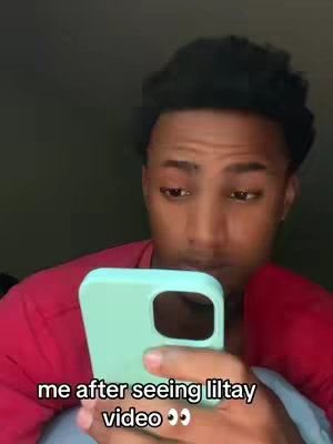 A young man, with short, textured black hair, is shown in a close-up view. He’s holding a phone, a mint green case surrounding it, in front of his face, but looking down at it. The lighting is dim, casting shadows and accentuating his features, and he wears a red shirt. The background is dark and out of focus.