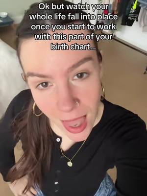 A selfie-style video with a young woman as the main subject, her face filling most of the frame. She has dark hair pulled back, a nose ring, and is wearing a black top with a gold necklace. Her mouth is open, tongue visible, and she appears to be speaking. The background is a bit blurry, showing a portion of a room with a cabinet.